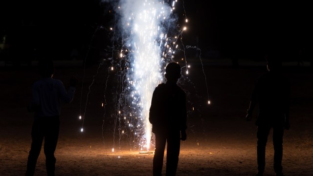 Three children silhouetted against a bright fountain firework illuminating a dark outdoor setting.