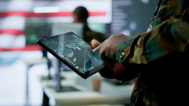 A person in military camouflage interacts with a tablet displaying a tactical map in a command center.