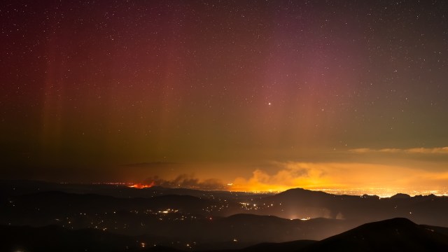A night sky filled with stars above smoky mountains and distant city lights, with a colorful aurora-like glow.
