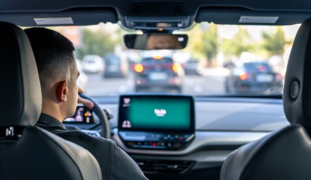 A driver is seated in a car, facing traffic with a digital dashboard display visible in front.
