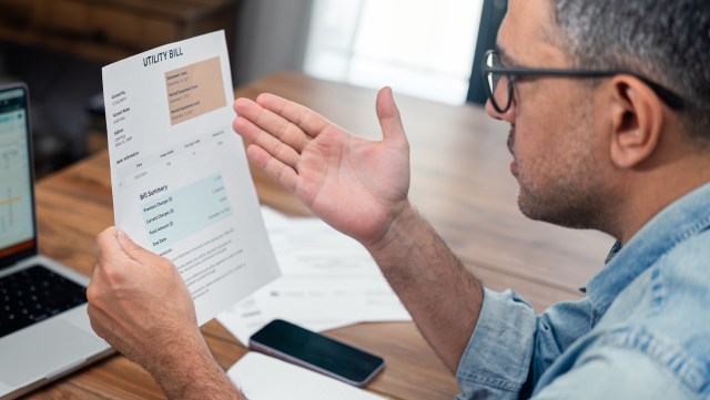 A man reviews a utility bill with a concerned expression while sitting at a desk with a laptop and smartphone.