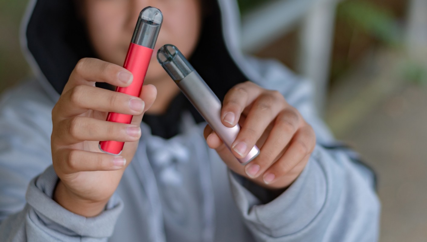A person holds two vaping devices, one red and one silver, in front of them.