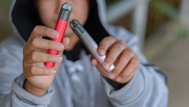 A person holds two vaping devices, one red and one silver, in front of them.