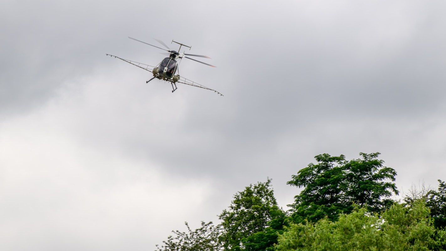 A helicopter flying above lush green trees under a cloudy sky.