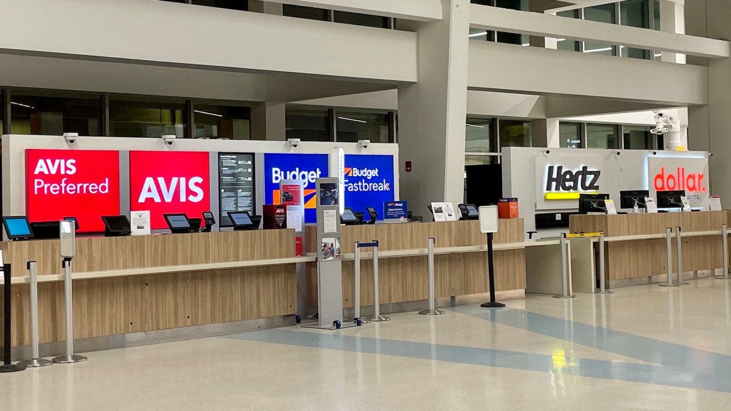 A car rental area at an airport displaying counters for Avis, Budget, Hertz, and Dollar.