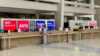 A car rental area at an airport displaying counters for Avis, Budget, Hertz, and Dollar.
