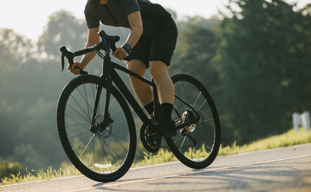A cyclist in athletic gear rides a black bike on a sunlit road surrounded by greenery.