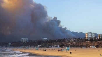 A beach scene with a distant plume of smoke rising from a hillside, under a clear sky at sunset.