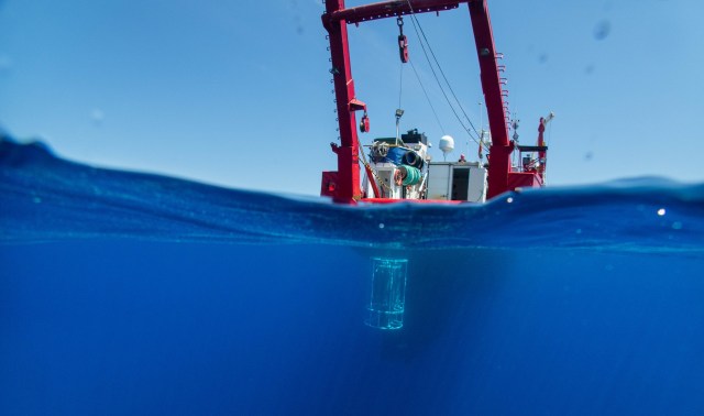 A research vessel is seen above water with a sampling device submerged below, under clear blue skies.