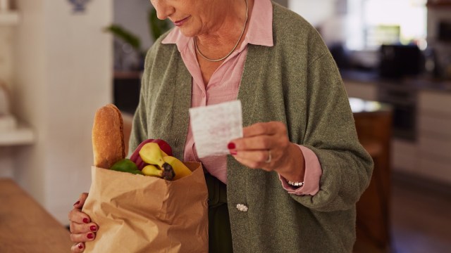 A woman examines a shopping list while holding a paper bag filled with fruits and a loaf of bread.