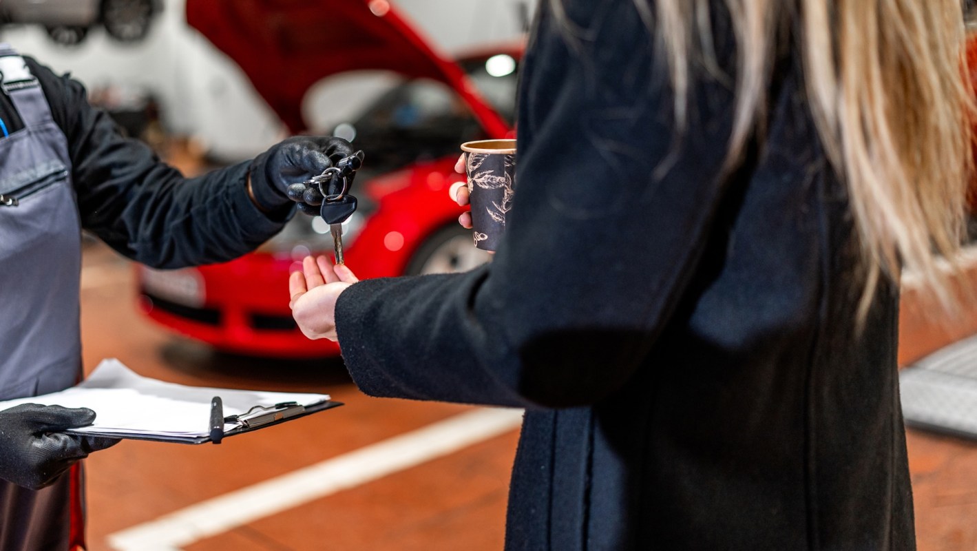 A mechanic hands keys to a woman while she holds a coffee cup in an automotive workshop.