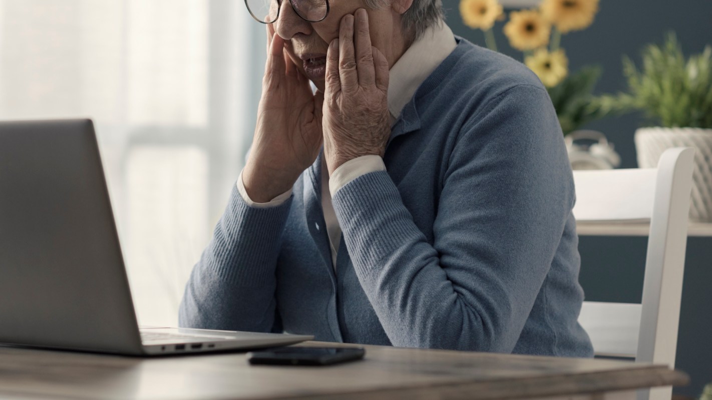 A senior woman with glasses looks at a laptop with a stressed expression.