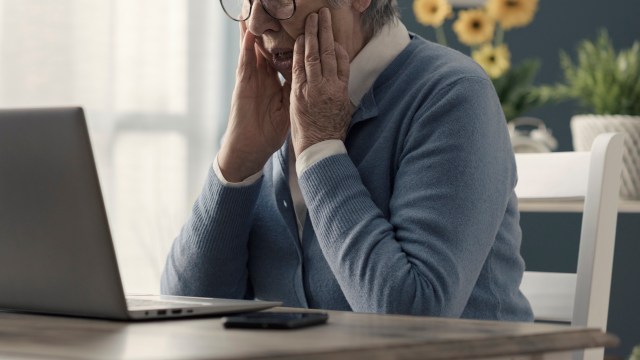 A senior woman with glasses looks at a laptop with a stressed expression.