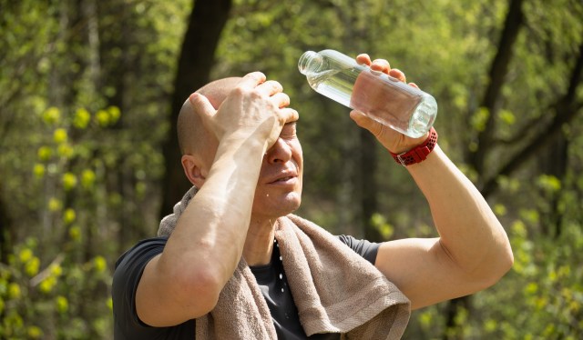 A person wipes sweat from their forehead while holding a water bottle in a wooded area.