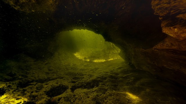 An underwater cave illuminated by a greenish light, showing swirling water and rocky textures.