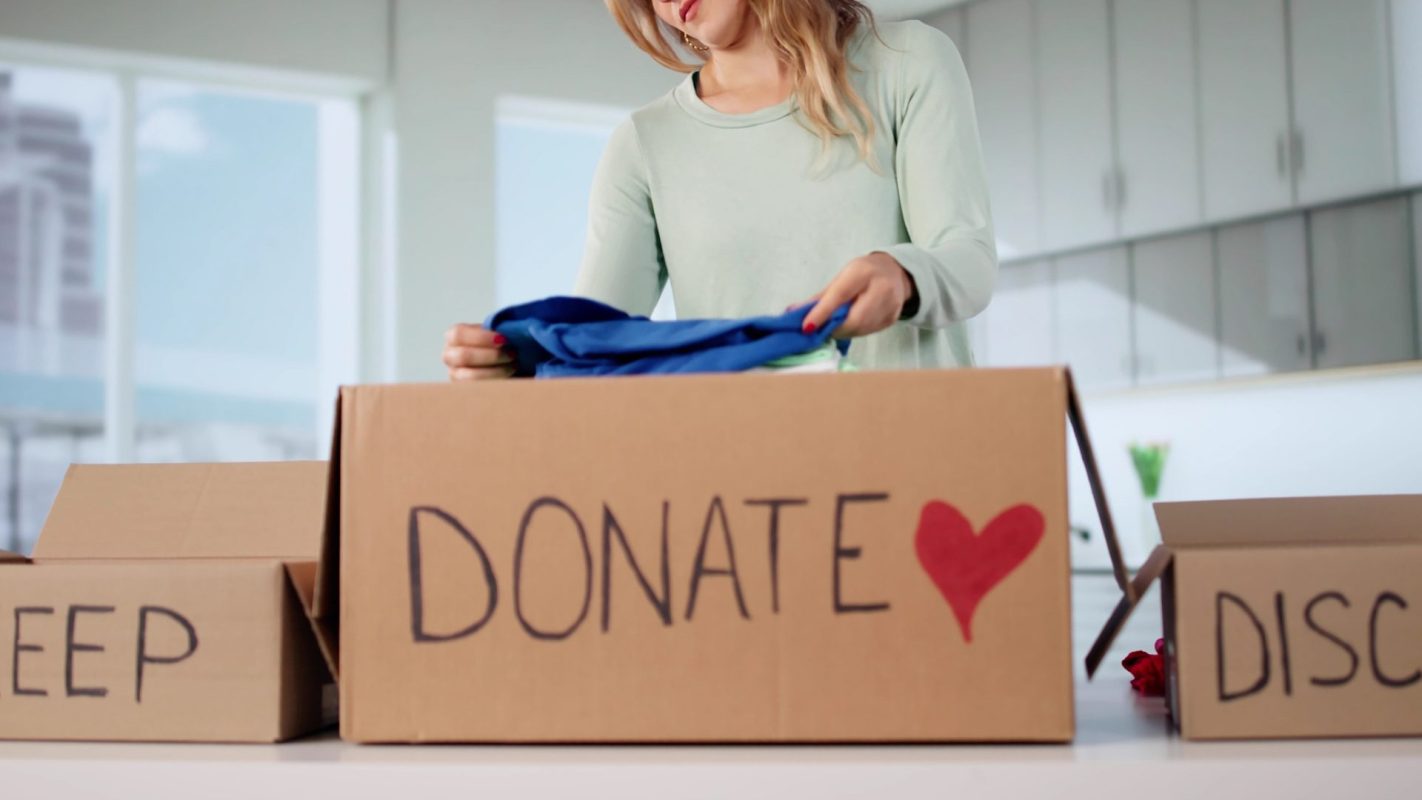 A woman places clothes into a cardboard box labeled 'DONATE' in a bright, modern room, with a 'KEEP' box nearby.