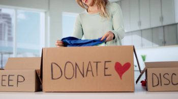 A woman places clothes into a cardboard box labeled 'DONATE' in a bright, modern room, with a 'KEEP' box nearby.