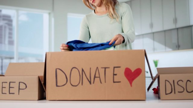 A woman places clothes into a cardboard box labeled 'DONATE' in a bright, modern room, with a 'KEEP' box nearby.