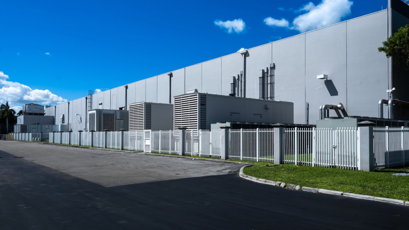 A large industrial building with mechanical units and a white fence under a clear blue sky.