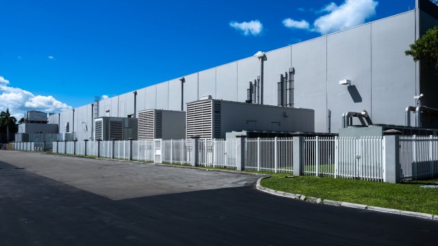 A large industrial building with mechanical units and a white fence under a clear blue sky.