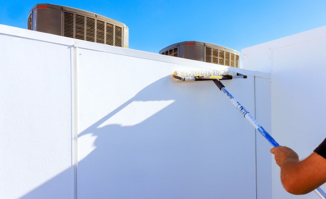 A person painting a white wall with a roller, near air conditioning units under a clear blue sky.