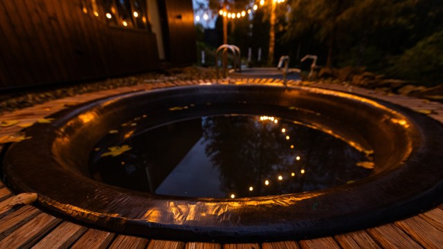 A round hot tub surrounded by wooden decking, with reflections of warm lights and autumn leaves floating on the water.