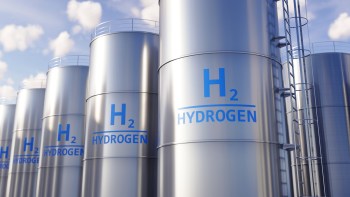 Row of metallic hydrogen storage tanks with blue "H2" and "HYDROGEN" labels against a cloudy sky.