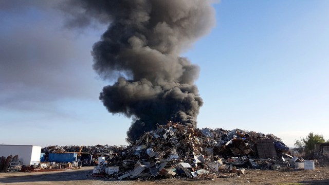 A large plume of black smoke rises over a scrap metal yard filled with piles of metal debris.