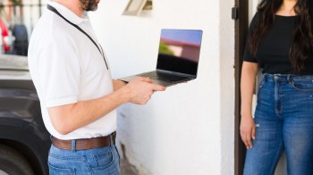 A man in a white polo shirt holds a laptop while talking to a woman in jeans at a doorway.