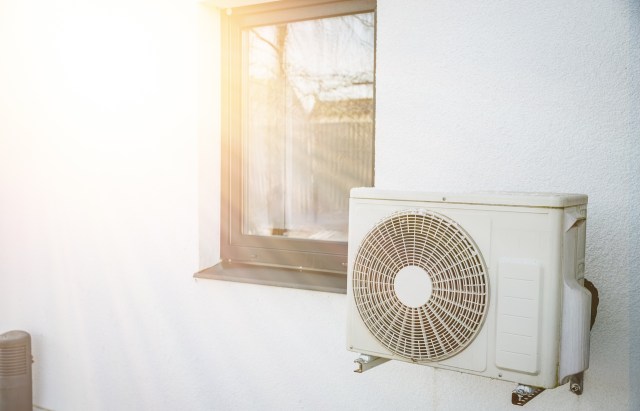 A heat pump unit mounted on a wall next to a window, illuminated by sunlight.