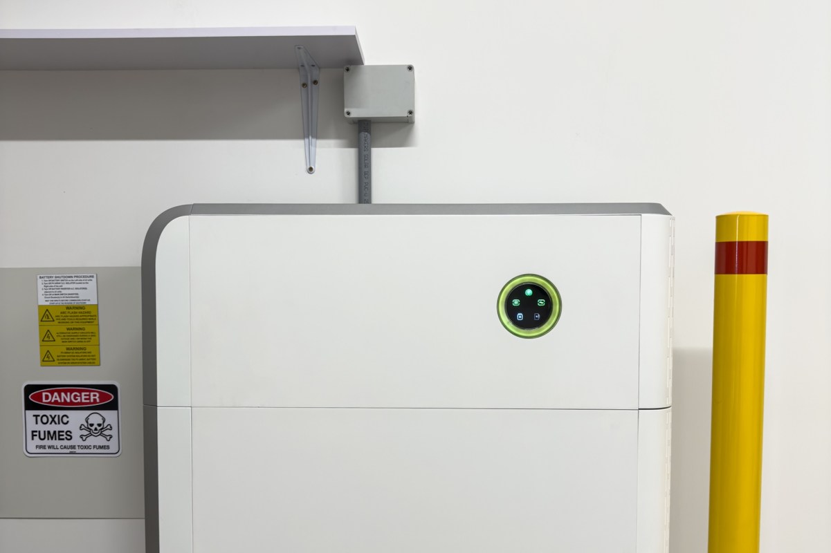 A white industrial battery storage unit on the wall beside a hazard warning.