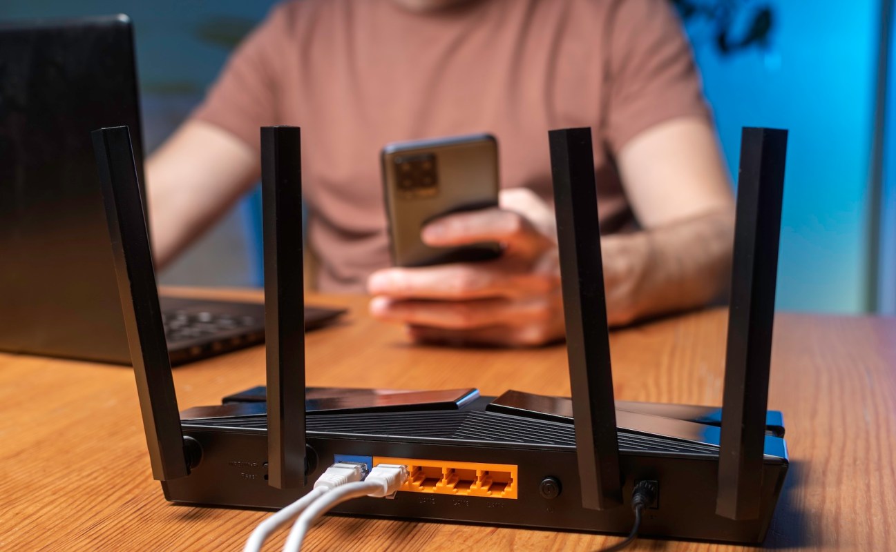 A Wi-Fi router lies on a wooden table as a person uses a smartphone in the background.