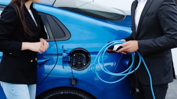 A man in a suit holds an electric vehicle charging cable while a woman in a blazer stands beside a blue car.