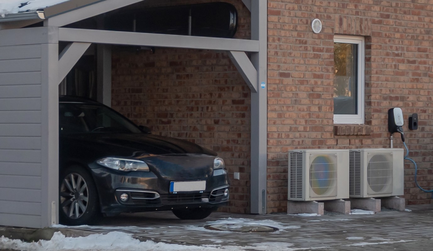 A black car in a carport near two AC units and an electric vehicle charging station.