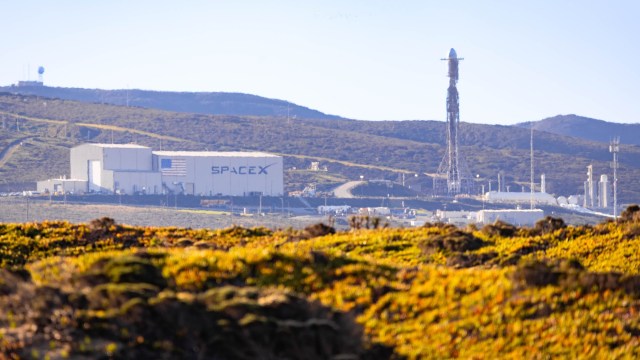 A SpaceX facility with a rocket and mountains in the background, surrounded by vibrant greenery.