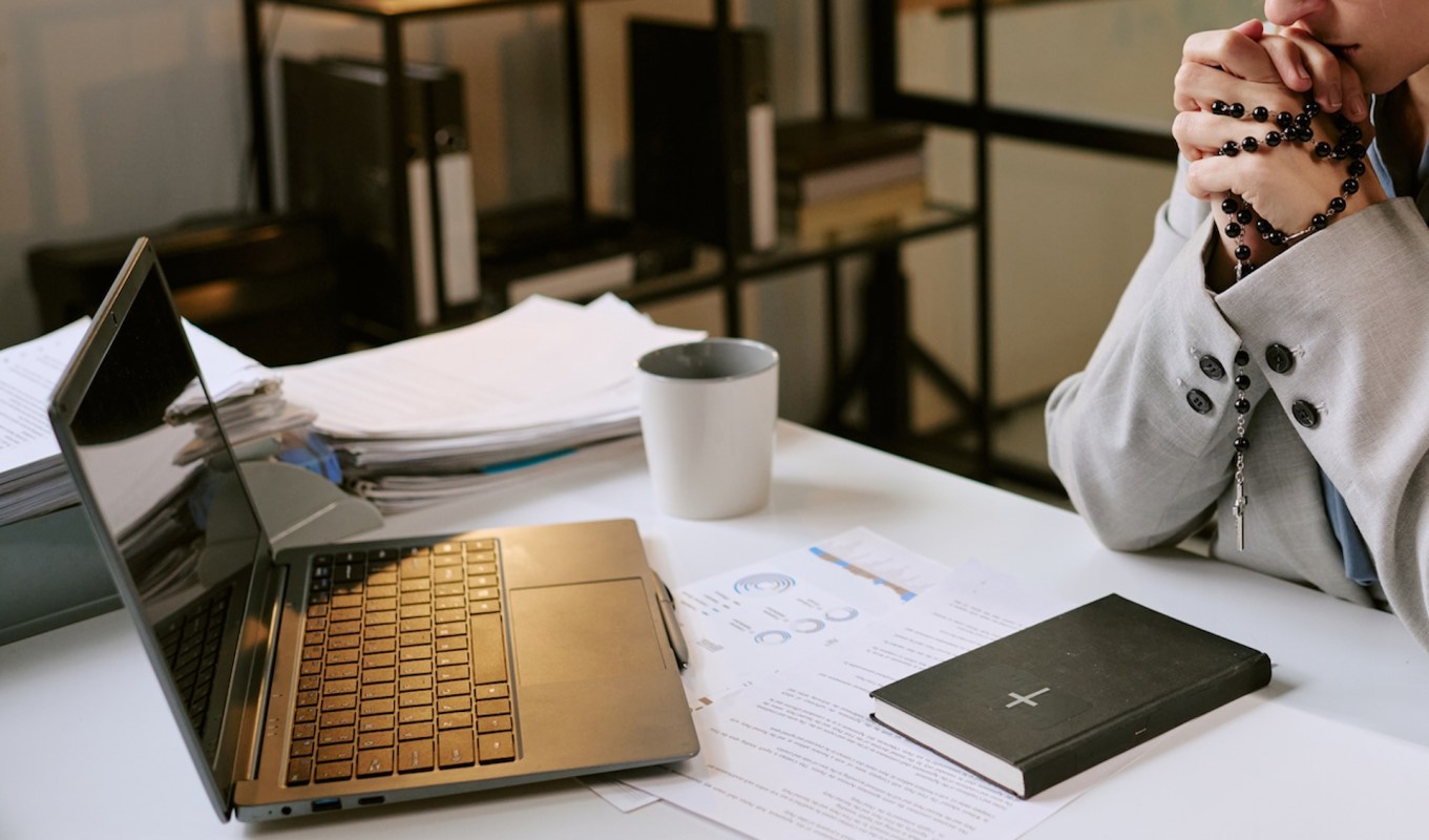 A person in a suit clasps their hands with a rosary beside a laptop and paperwork on a table.