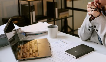 A person in a suit clasps their hands with a rosary beside a laptop and paperwork on a table.