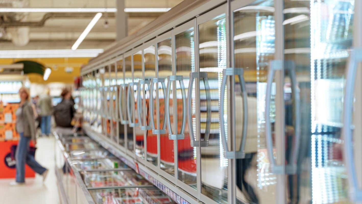 A row of glass-fronted freezer cases in a grocery store with people shopping in the background.