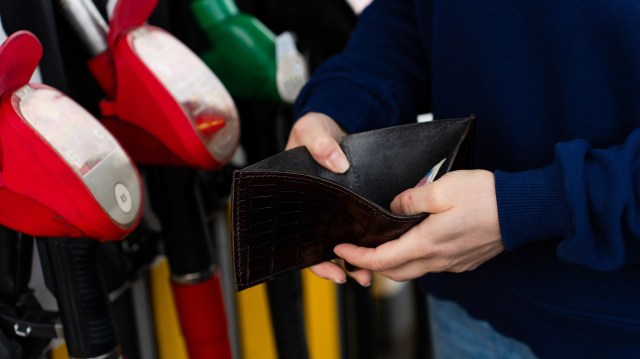 A person holds an open wallet while standing in front of gas pumps at a fuel station.