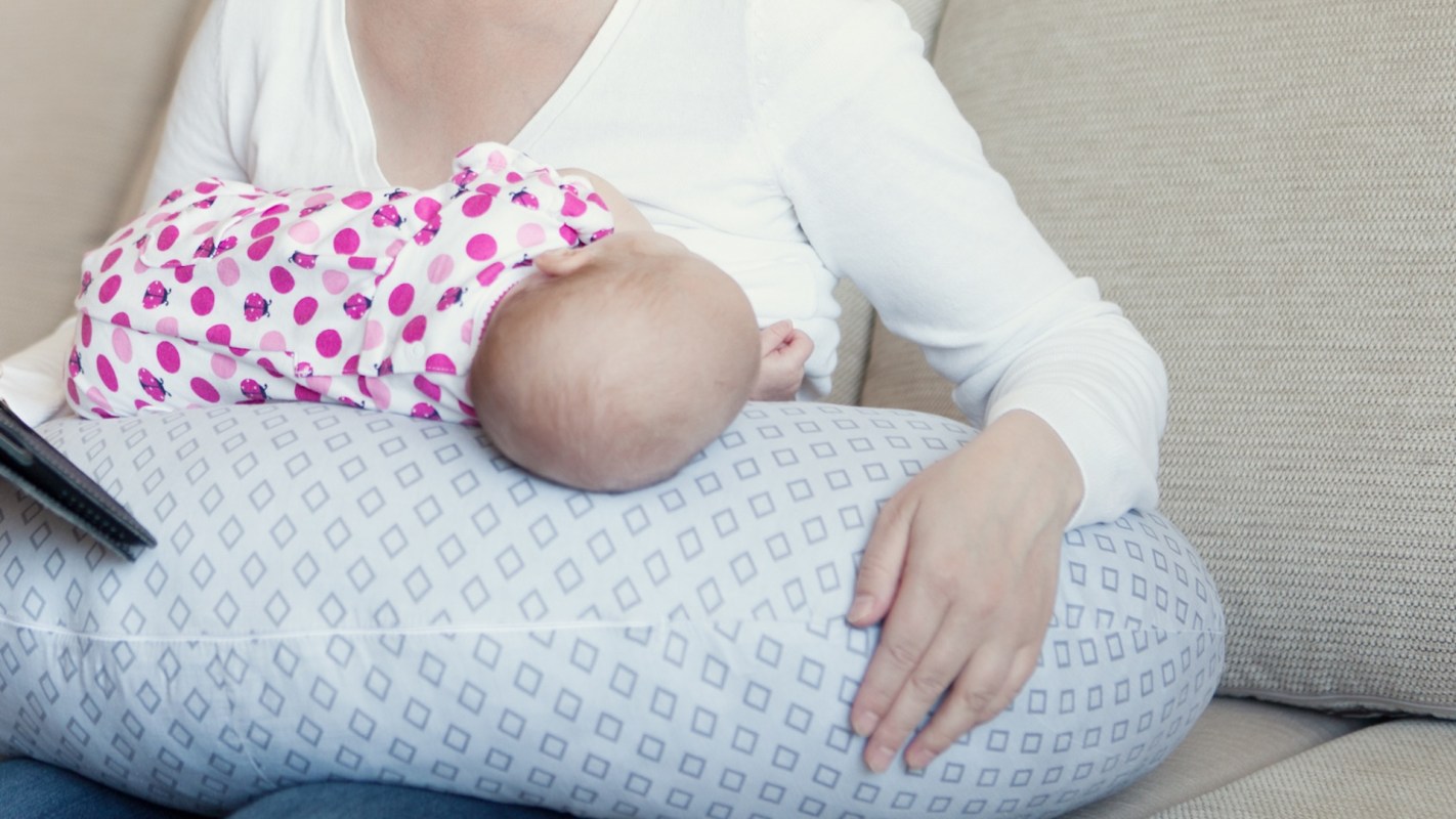 A mother sitting on a couch, breastfeeding a baby while holding a tablet.