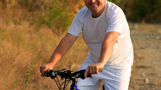 A smiling man in a white T-shirt riding a mountain bike on a dirt path surrounded by greenery.