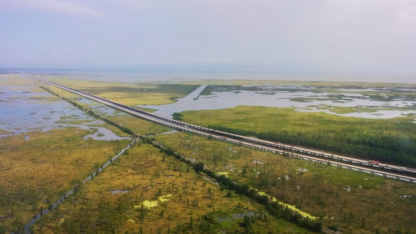 An aerial view of a long highway bordered by wetlands and patches of greenery under a cloudy sky.