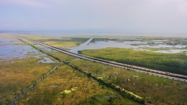 An aerial view of a long highway bordered by wetlands and patches of greenery under a cloudy sky.
