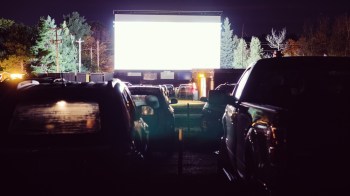 A dimly lit drive-in theater with cars parked in front of a large blank screen at night.