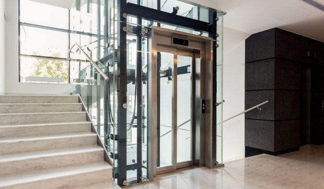 A modern interior featuring a glass elevator beside a marble staircase in a well-lit building.