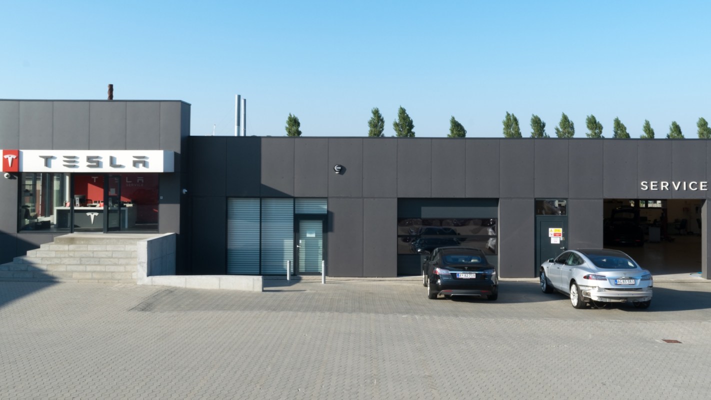A Tesla service center displaying three parked vehicles and a prominent Tesla logo on the building.