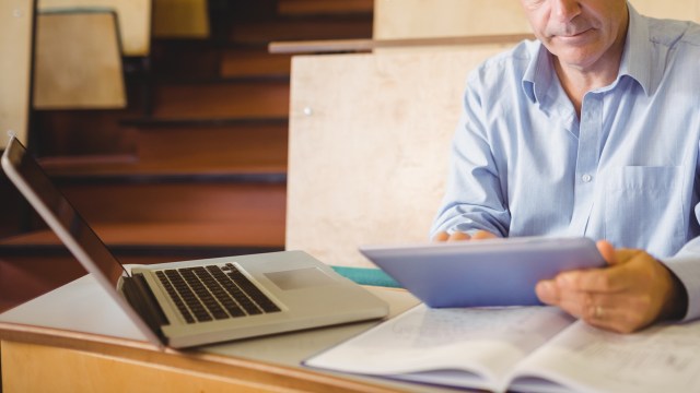 A professor in a blue shirt examines a tablet while sitting at a desk with a laptop and an open notebook.