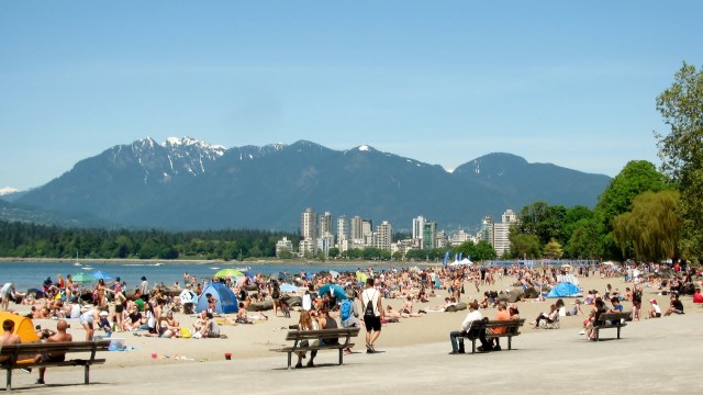 A crowded beach with people sunbathing, playing, and enjoying the view of mountains and city skyline on a sunny day.