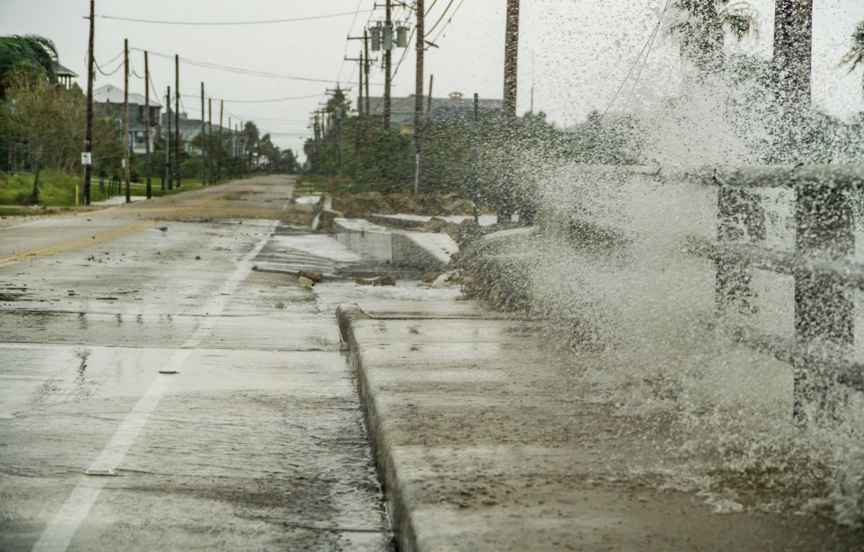 A flooded street with water splashing against a concrete barrier, amidst a gray, overcast sky.