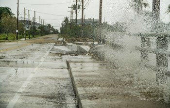 A flooded street with water splashing against a concrete barrier, amidst a gray, overcast sky.